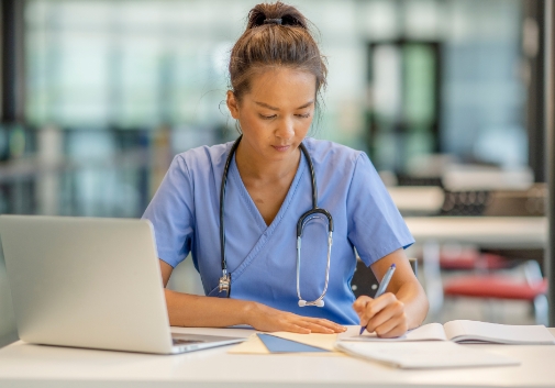 A healthcare support professional writing notes in a notepad while sitting at a desk with a laptop and papers 