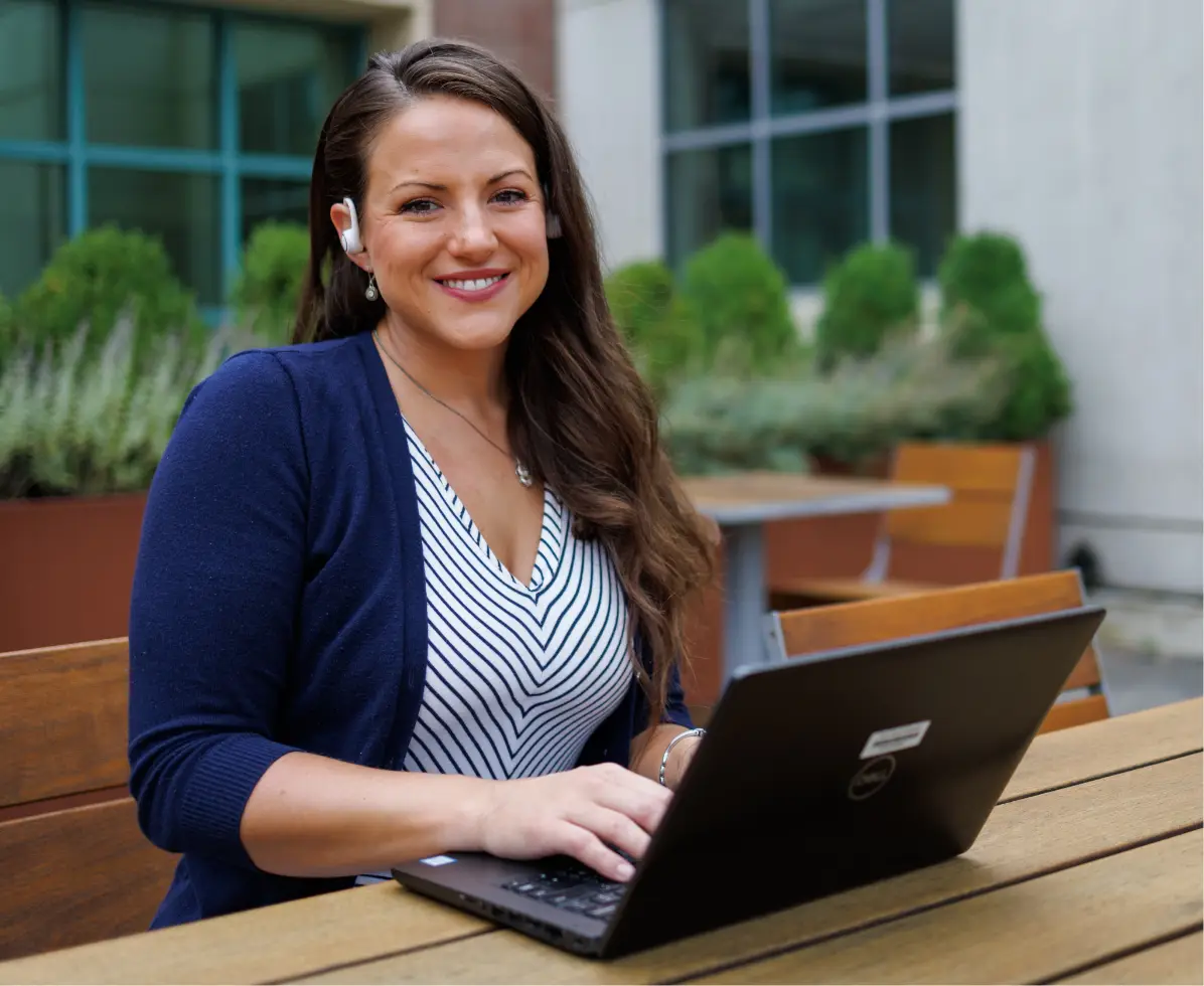 A woman smiling while working on a laptop outside.